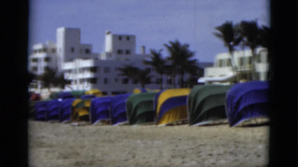 1975 Florida beach footage frame with tent rows, white seaside buildings, palm trees, a small white vehicle, and sand in front.