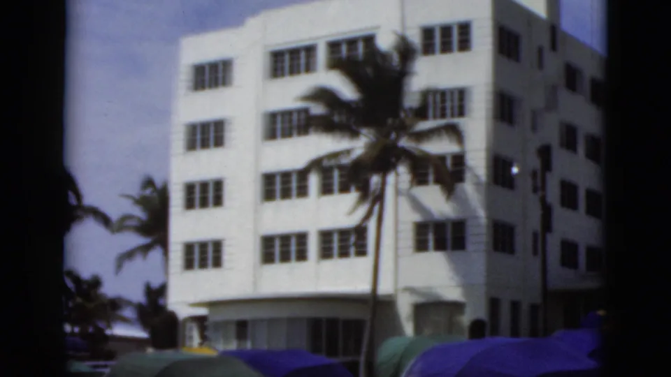 1975 Florida beach view with a white multi-story building, palm tree, and colorful beach shades in the foreground.