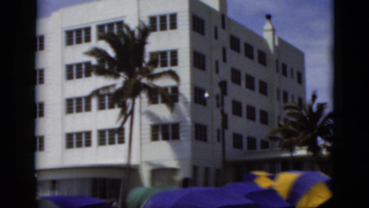 Colorful umbrellas and canvas beach tents sit before a white Florida beach building with palms nearby.