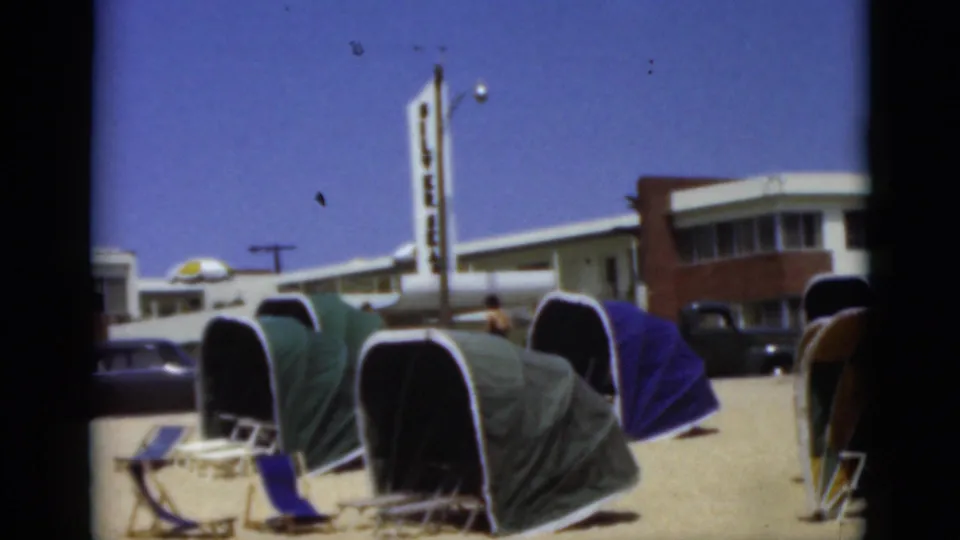 Florida beach cabanas overlook a busy resort street with parked vintage cars, a low motel-like building, and a tall sign.