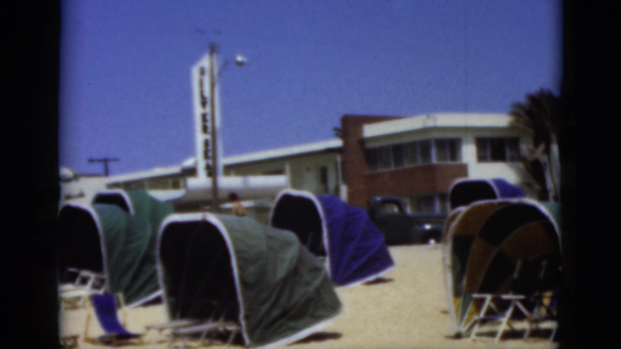 1975 Florida 8mm frame showing retro dome beach tents on sand with a low beachside building, palm trees, tall sign, and parked dark vehicle behind them.