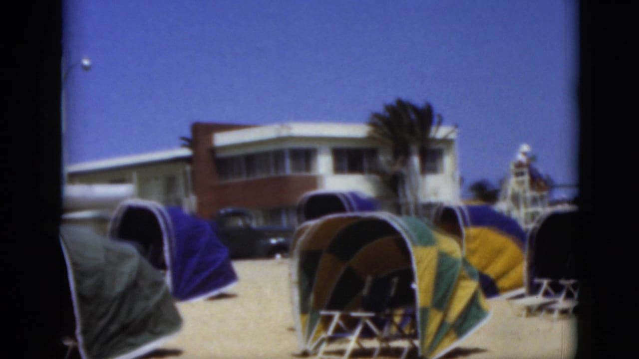 1975 Florida beach view with clustered retro dome tents, a low modern building, palm tree, dark vehicle, and lifeguard tower under a clear sky.