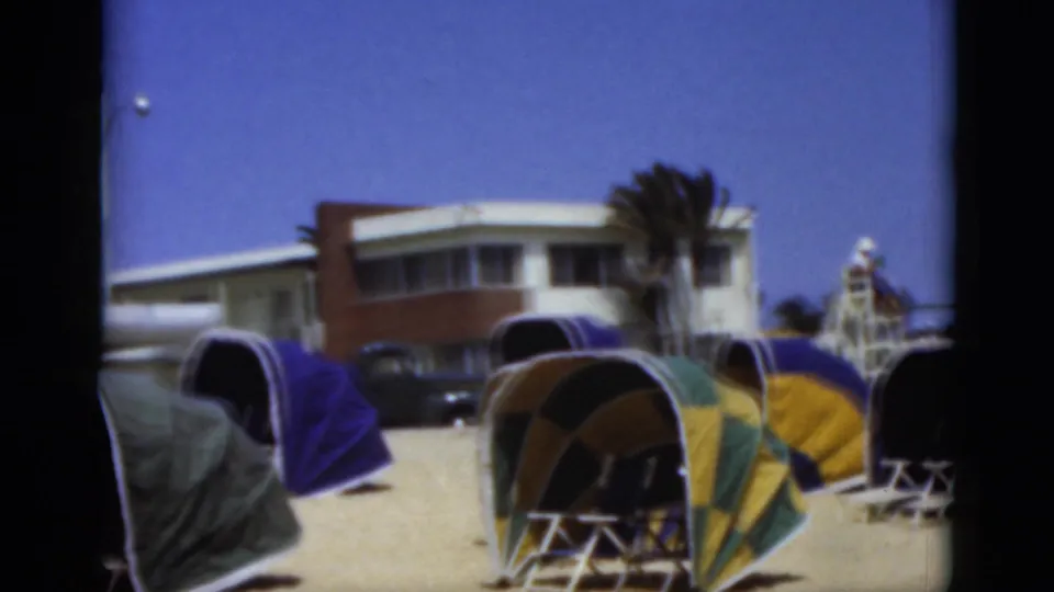 1975 Florida beach view with clustered retro dome tents, a low modern building, palm tree, dark vehicle, and lifeguard tower under a clear sky.