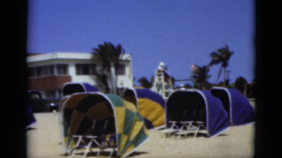 1975 Florida home-movie frame showing multiple dome beach shelters on sand, with palm trees, a lifeguard tower, and a beachside building behind them.