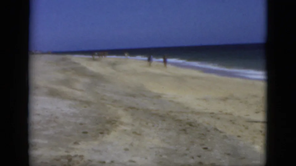 1975 Florida shoreline view on 8mm film, with open sand, ocean surf, a blue horizon, and small distant beachgoers near the waterline.