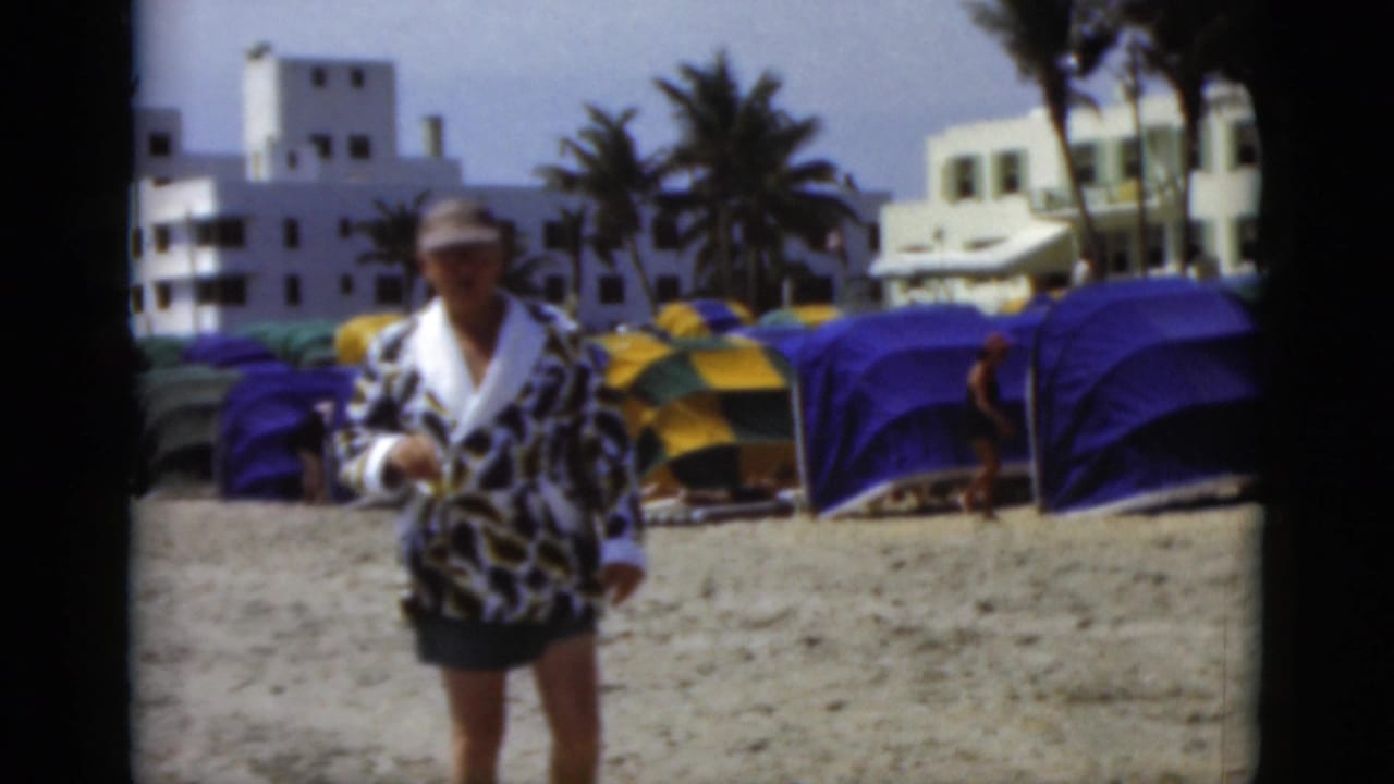 Man in patterned robe and cap walking on a sandy beach near colorful cabanas.