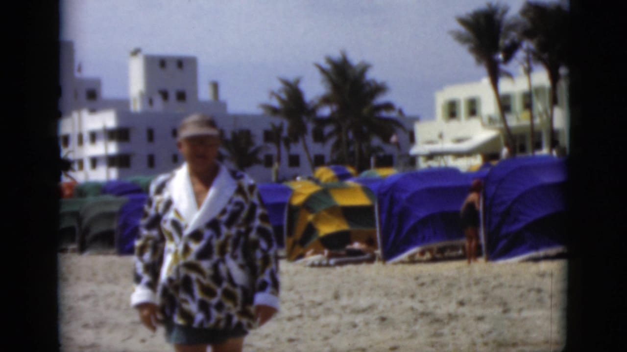 The man in a bold robe and cap appears closer to the camera, with colorful cabanas, palms, and white coastal buildings behind him.