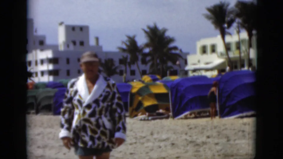 The man in a bold robe and cap appears closer to the camera, with colorful cabanas, palms, and white coastal buildings behind him.