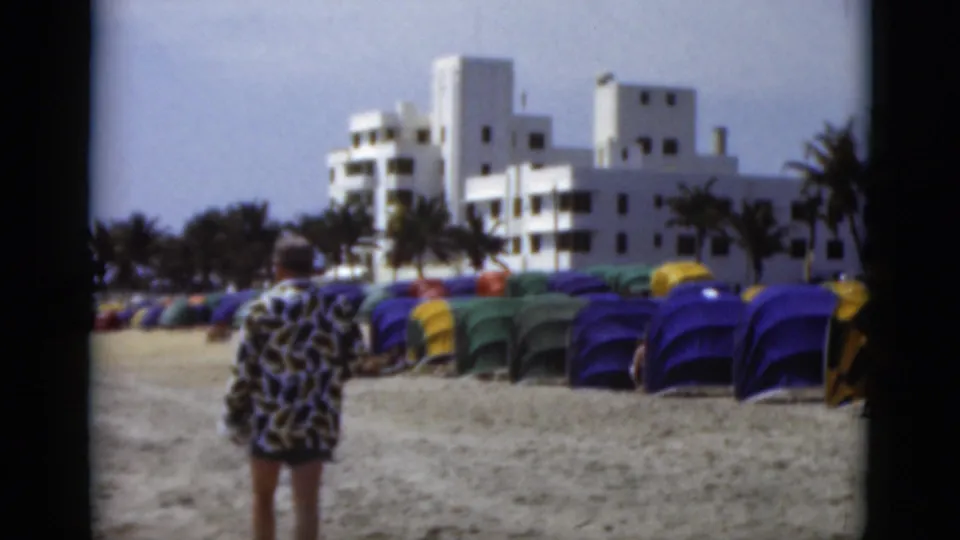 Viewed from behind, the man in the patterned robe walks across the sand toward rows of cabanas and white buildings in Florida beach footage.