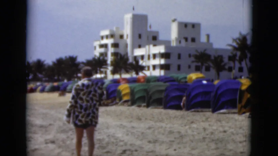The final frame shows the man from behind on the sand, facing rows of blue, green, and yellow cabanas below white coastal buildings.