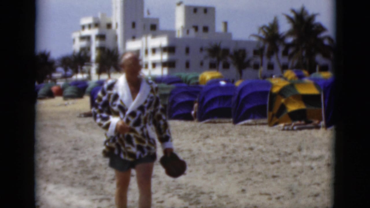 A man in a leopard-print robe approaches the camera on a Florida beach, with colorful tents, palm trees, and a white building behind him.
