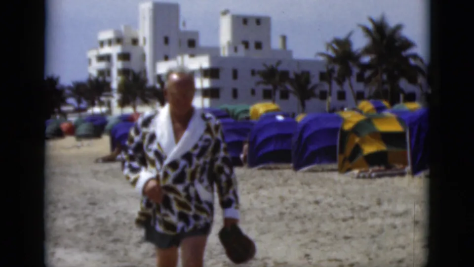The man appears closer in soft-focus 8mm footage, standing on a Florida beach before rows of blue and yellow tents and palm trees.