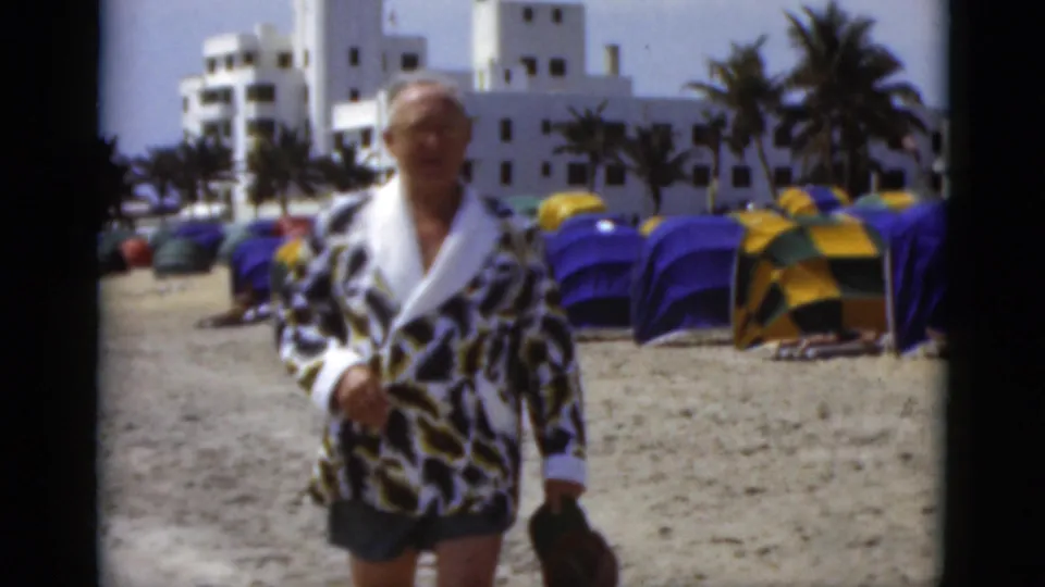 A man in a leopard-print robe remains in the foreground of a 1975 Florida beach scene, with tent rows, palms, and a white resort-like building visible.