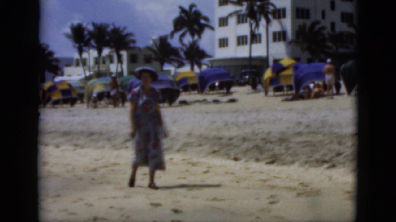 Woman in a floral dress and hat walking on a sandy beach with umbrellas and buildings behind her.