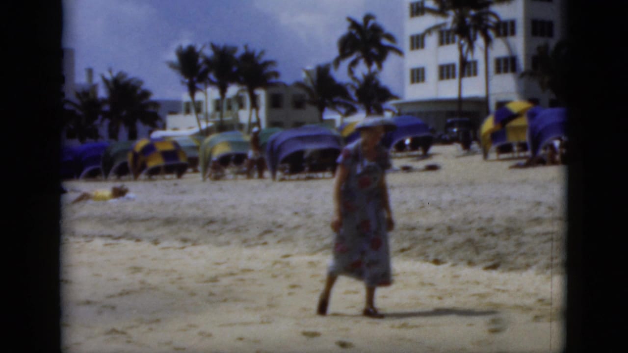 Woman walking on sand near beach umbrellas, palm trees, and white buildings.