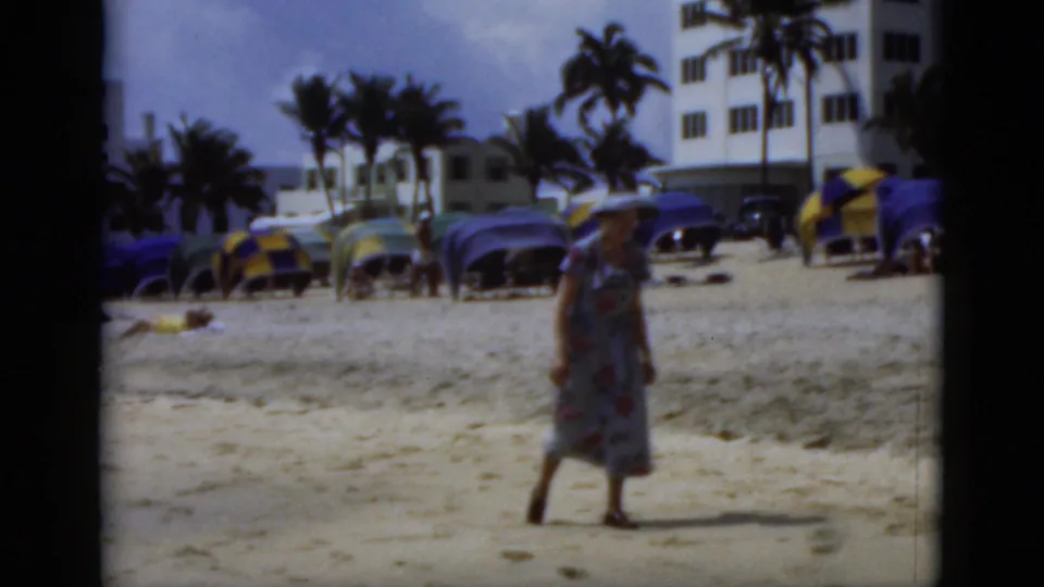 1975 Florida beach scene showing the woman farther along the sand, with rows of umbrellas, palms, and shoreline buildings behind her.