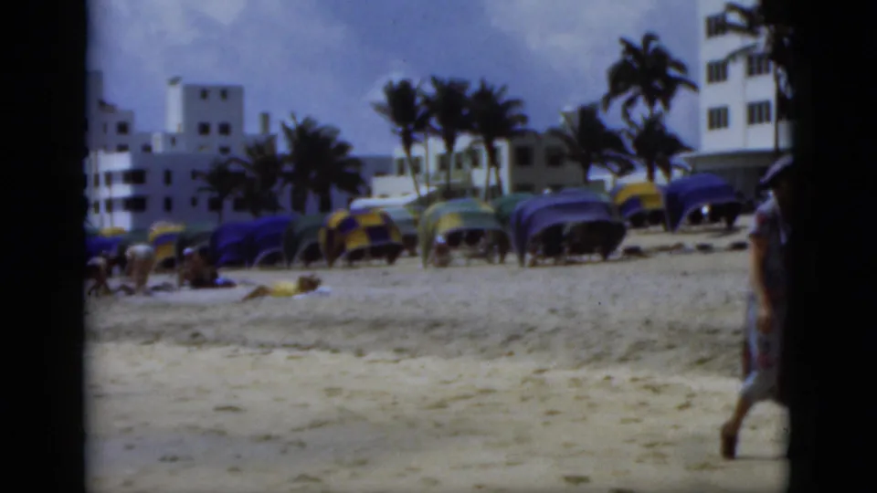 1975 Florida home-movie frame centered on beach umbrellas, palms, and white buildings, with the walking woman partly cropped at right.