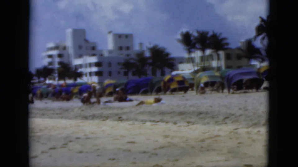 1975 Florida beach view with colorful umbrellas, scattered beachgoers, palm trees, and white buildings along the shore.
