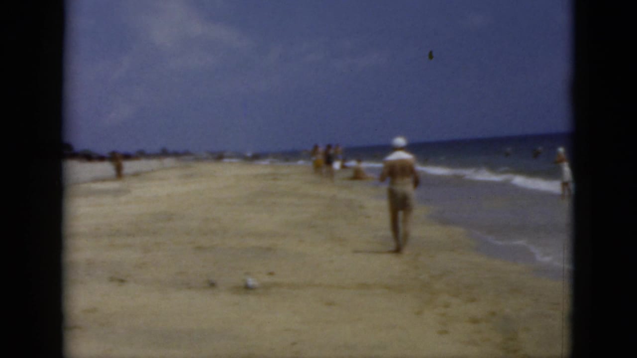 1975 Florida 8mm beach frame showing a man walking away near gentle waves under a cloudy sky.