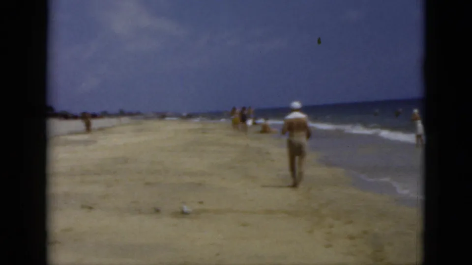 1975 Florida 8mm beach frame showing a man walking away near gentle waves under a cloudy sky.