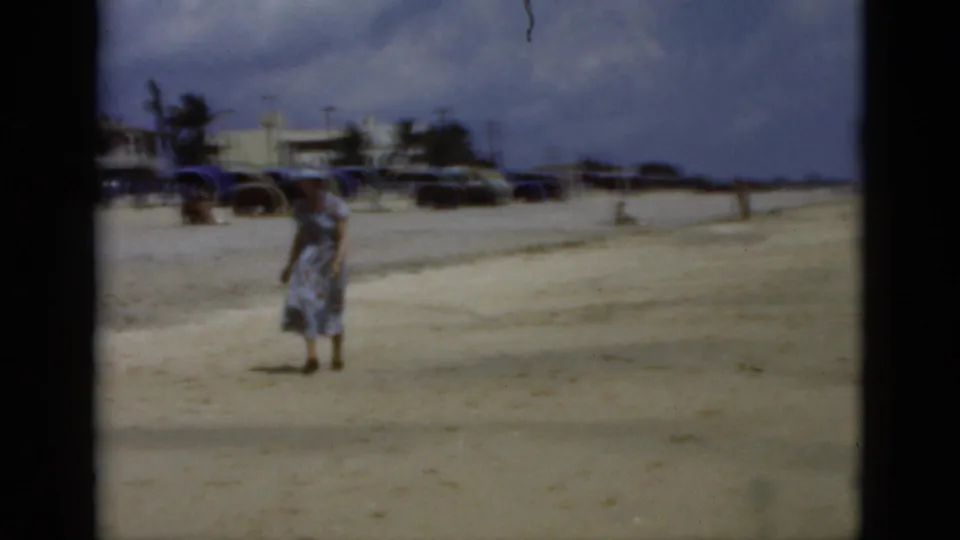 1975 Florida 8mm frame showing a woman bending on the beach, with cars, palms and umbrellas or cabanas in the background.