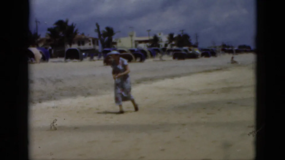1975 Florida beach frame with a woman near the foreground, parked cars, palms, cabanas or umbrellas and cloudy sky in view.