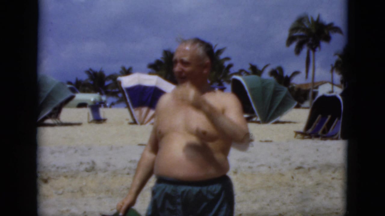 Shirtless older man in swim trunks standing on a sandy beach with umbrellas and palm trees behind him.