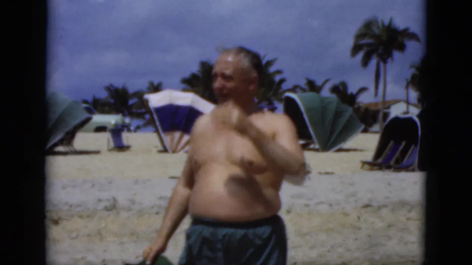 1975 Florida 8mm beach frame showing a shirtless older man in dark swim trunks near umbrellas, lounge chairs, sand, and palm trees.