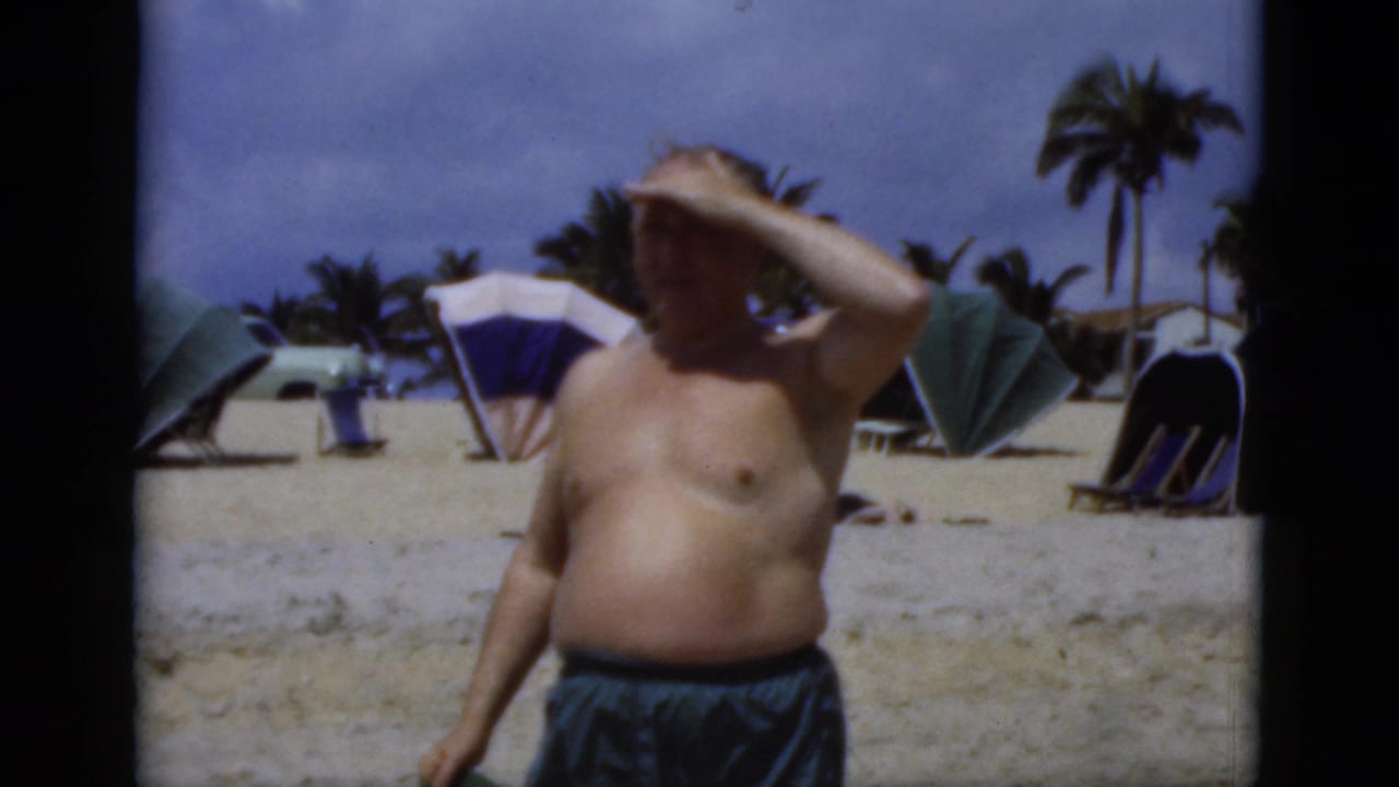 1975 Florida beach home-movie frame of an older man shielding his eyes from the sun near beach umbrellas and palm trees.