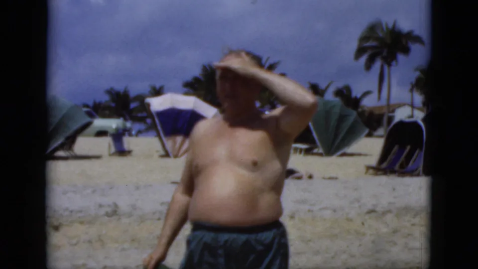 1975 Florida beach home-movie frame of an older man shielding his eyes from the sun near beach umbrellas and palm trees.