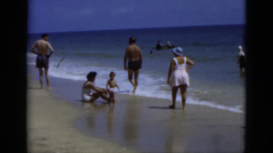 1975 Florida 8mm shoreline view with several beachgoers near the surf, including a woman in a white swimsuit and hat.
