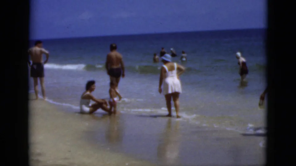 1975 Florida beach frame showing people in shallow surf, wet sand reflections, and a woman in a white swimsuit and hat.