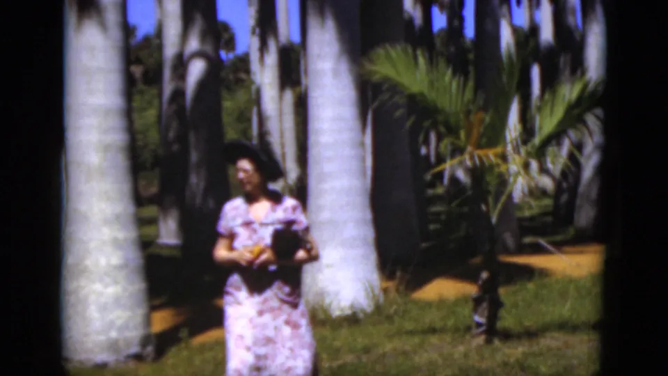 A woman in a floral dress and hat holds a small object while standing among palms in sunny 1975 Florida footage.