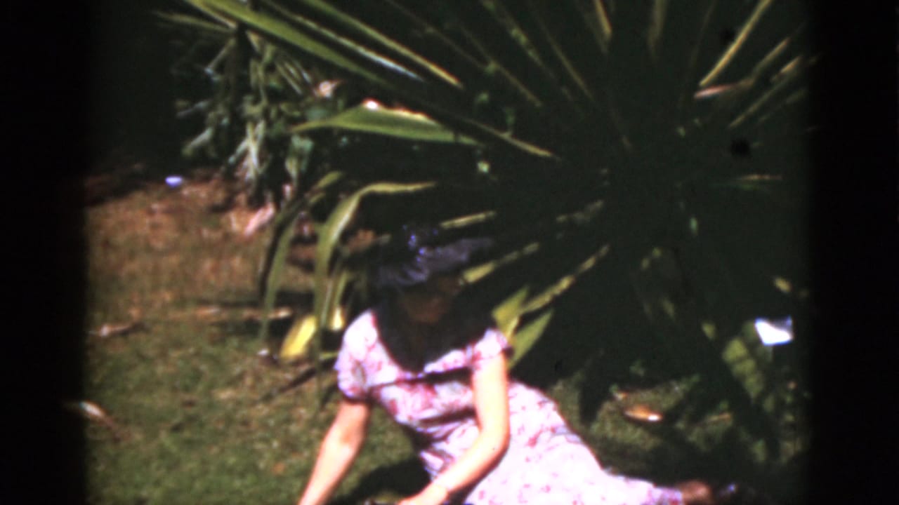 A woman in a purple floral dress and dark hat sits near large tropical plants in a Florida garden, 1975.