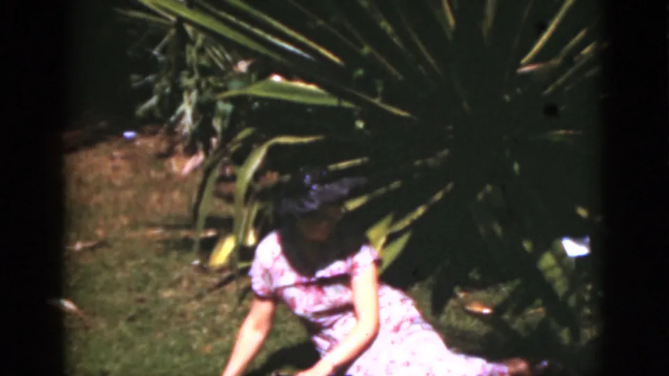 A woman in a purple floral dress and dark hat sits near large tropical plants in a Florida garden, 1975.