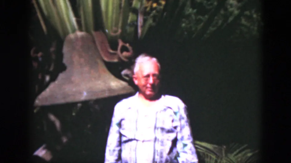Older man in a patterned shirt stands near a large weathered bell in the 1975 Florida home-movie footage.