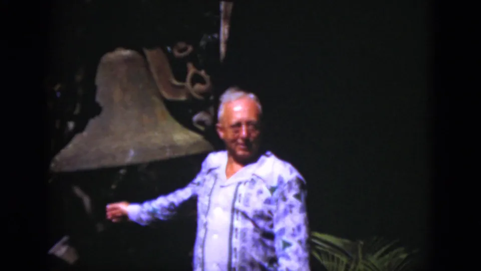 Smiling older man gestures toward a large bell in a shaded Florida outdoor scene recorded on 8mm film.