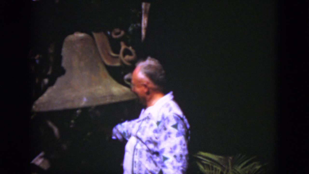 In 1975 Florida 8mm footage, an adult man in a blue patterned shirt stands near a large historic bell against a dark background.