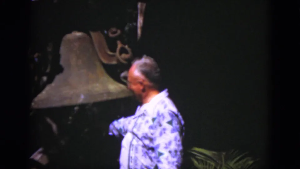 In 1975 Florida 8mm footage, an adult man in a blue patterned shirt stands near a large historic bell against a dark background.