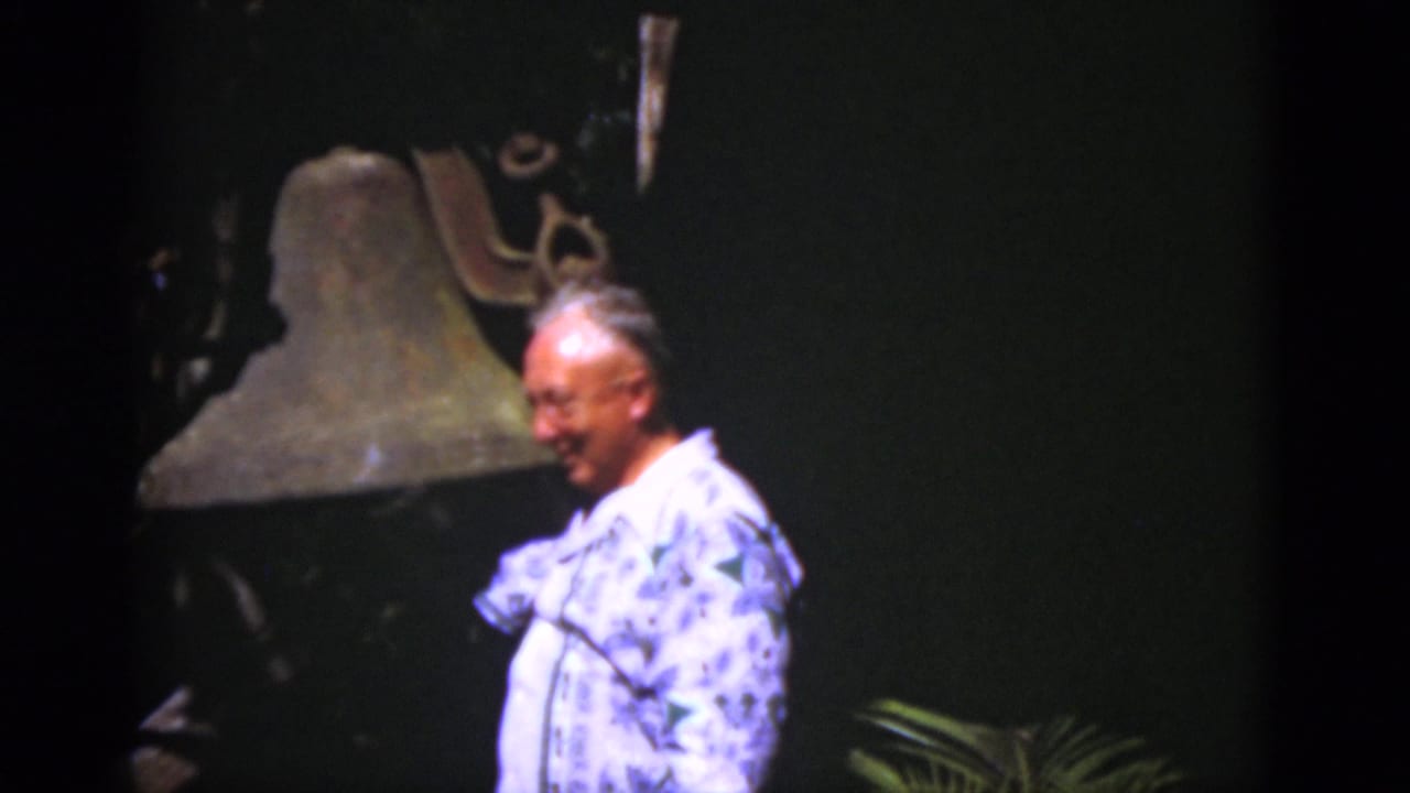 The man smiles and turns beside the large bell, with the dark backdrop and soft 8mm color suggesting a historic display.