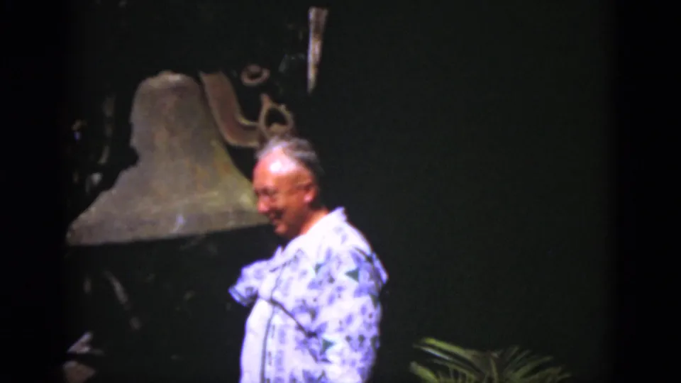 The man smiles and turns beside the large bell, with the dark backdrop and soft 8mm color suggesting a historic display.