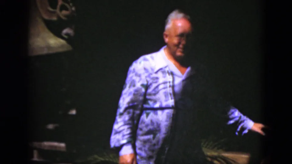 The man moves away from the bell area, with part of the metal bell still visible at left and a dark exhibit-like background behind him.