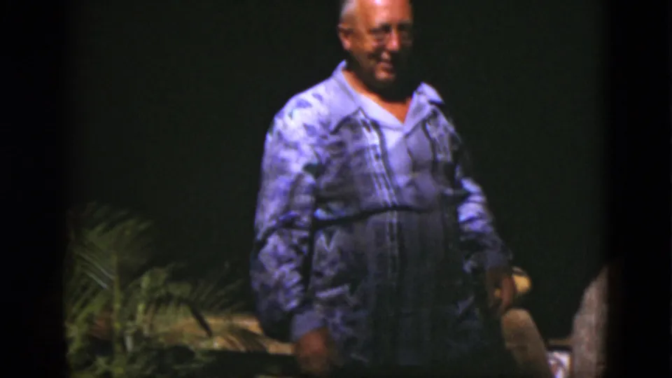 The man stands in front of a dark backdrop with leafy plants nearby, after moving past the large bell in the 1975 Florida footage.