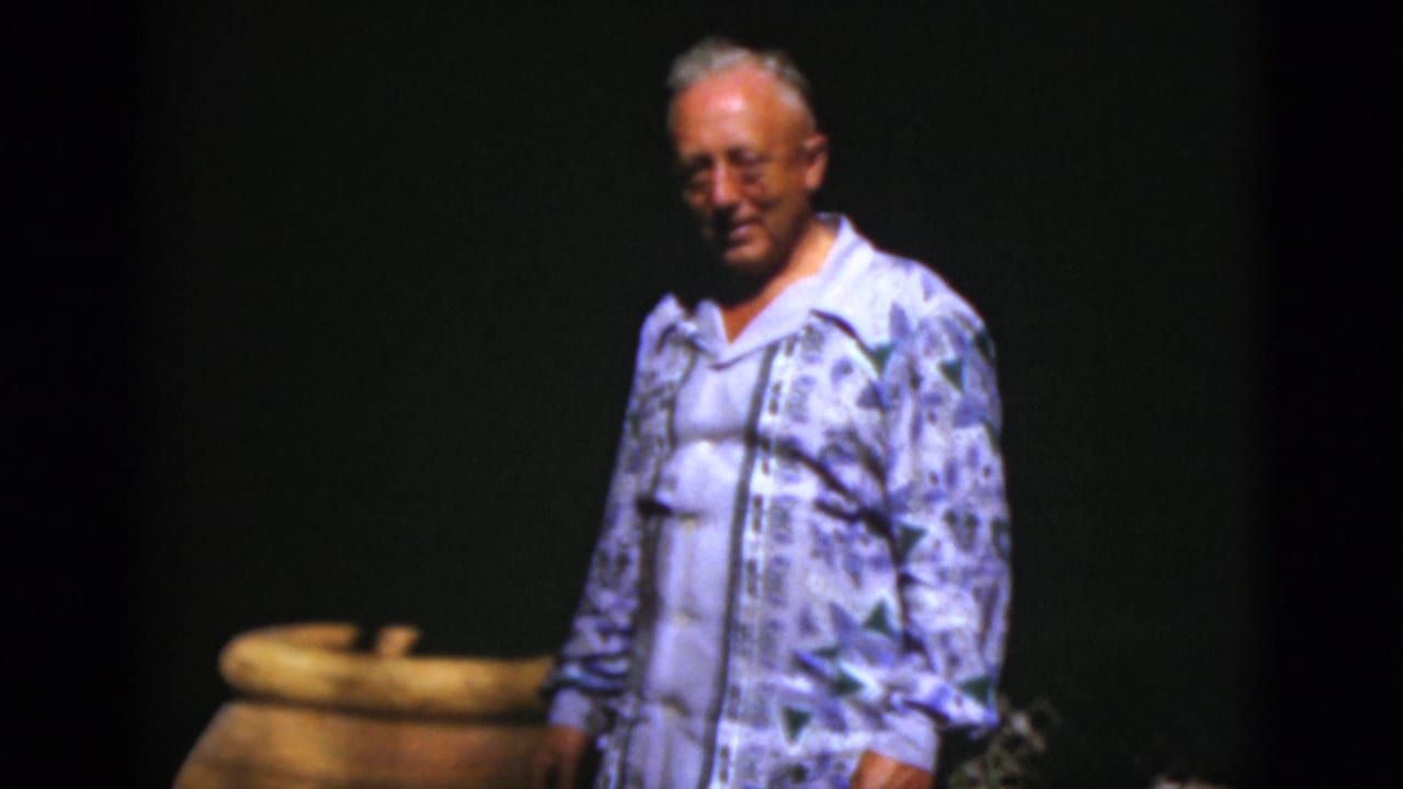 Older man in a patterned shirt faces the camera near a large pot.