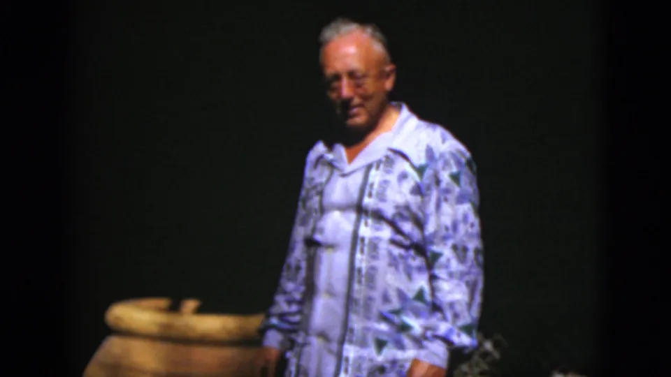 An older man in glasses and a light patterned shirt faces the camera beside a large ceramic pot in 1975 Florida home-movie footage.