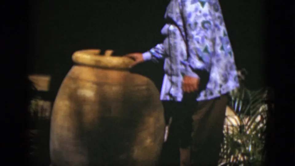 The man moves closer and reaches toward the rim of the large ceramic pot, with plants and a dark backdrop partly visible.