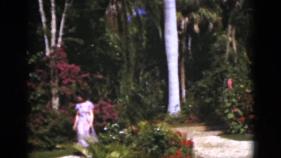 1975 Florida 8mm frame showing the woman in a pale pink dress crossing the garden lawn with palms, flowers, and a bright path nearby.