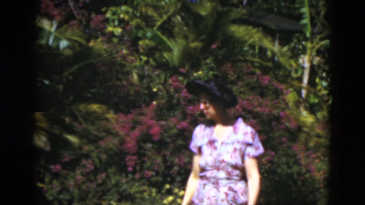 Woman in a floral dress and dark hat standing near flowering bushes and palm fronds.