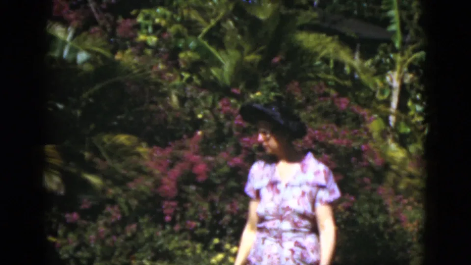 The woman appears closer to the camera in a Florida tropical garden, wearing a patterned dress and dark hat among bright foliage.