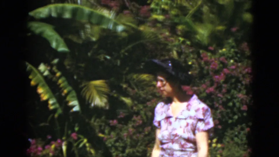 A close home-movie view shows the woman in a floral dress and dark hat beside tropical leaves and purple blossoms in Florida.